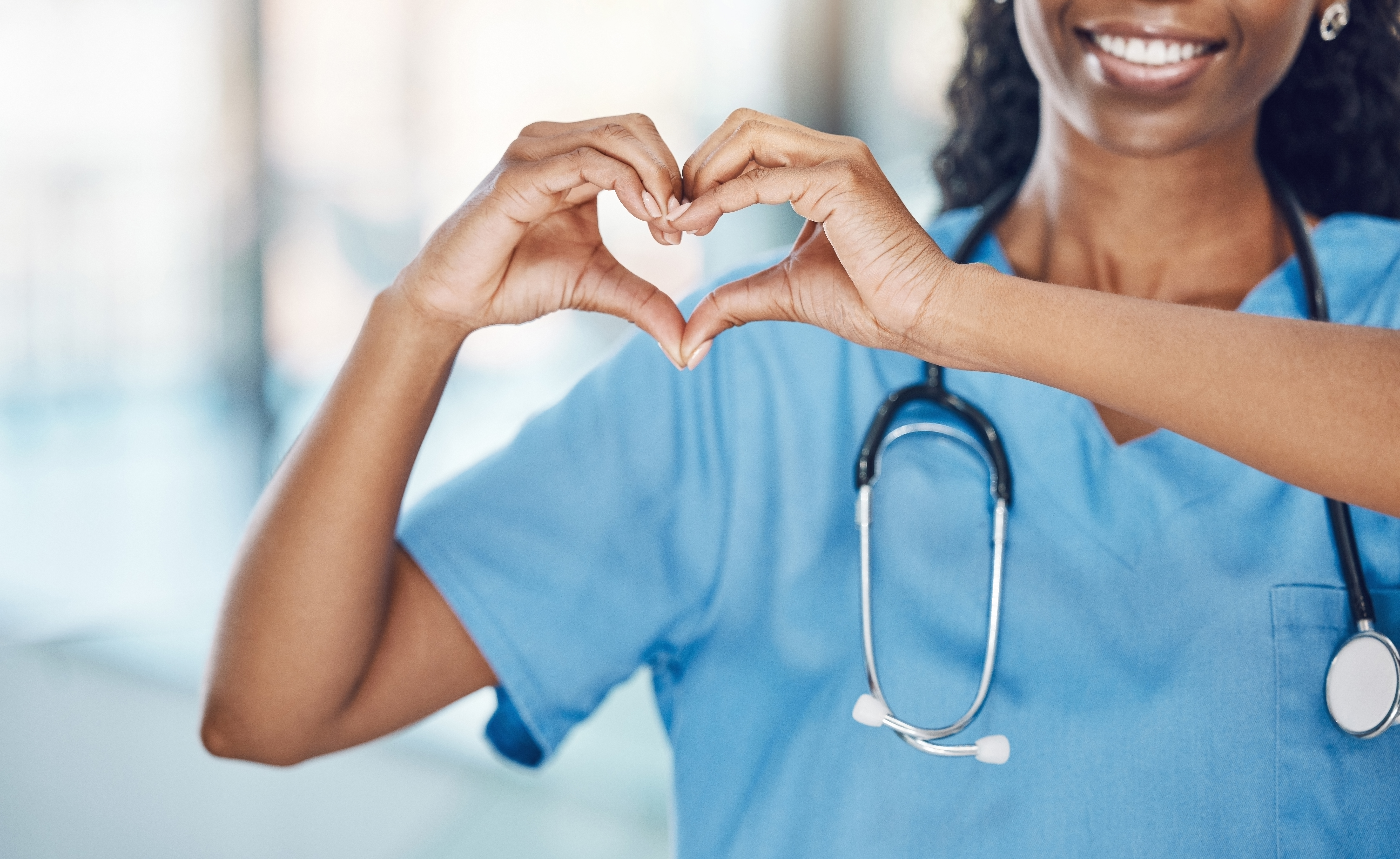 Nurse making a heart with her hands wearing blue scrubs with a stethoscope around her neck.