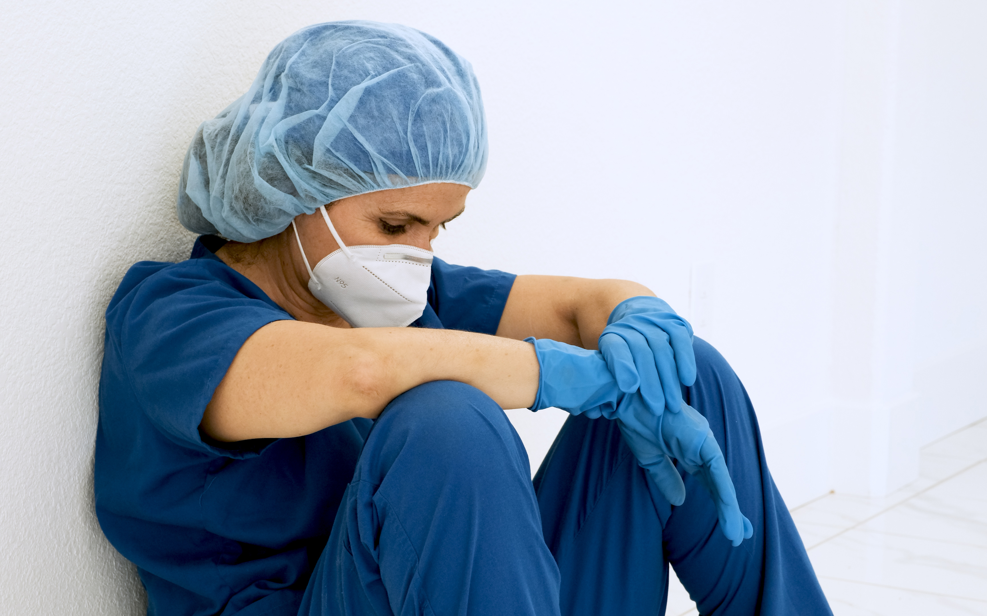 nurse in blue scrubs with hair net, face mask, and blue gloves. Nurse is sitting against a wall 