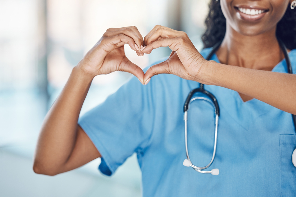 Nurse making a heart with her hands wearing blue scrubs with a stethoscope around her neck.