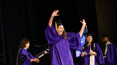 Graduating student in purple Regalia hands above head cheering in excitement