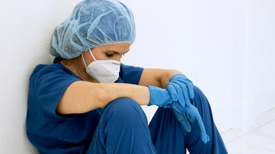 nurse in blue scrubs with hair net, face mask, and blue gloves. Nurse is sitting against a wall 